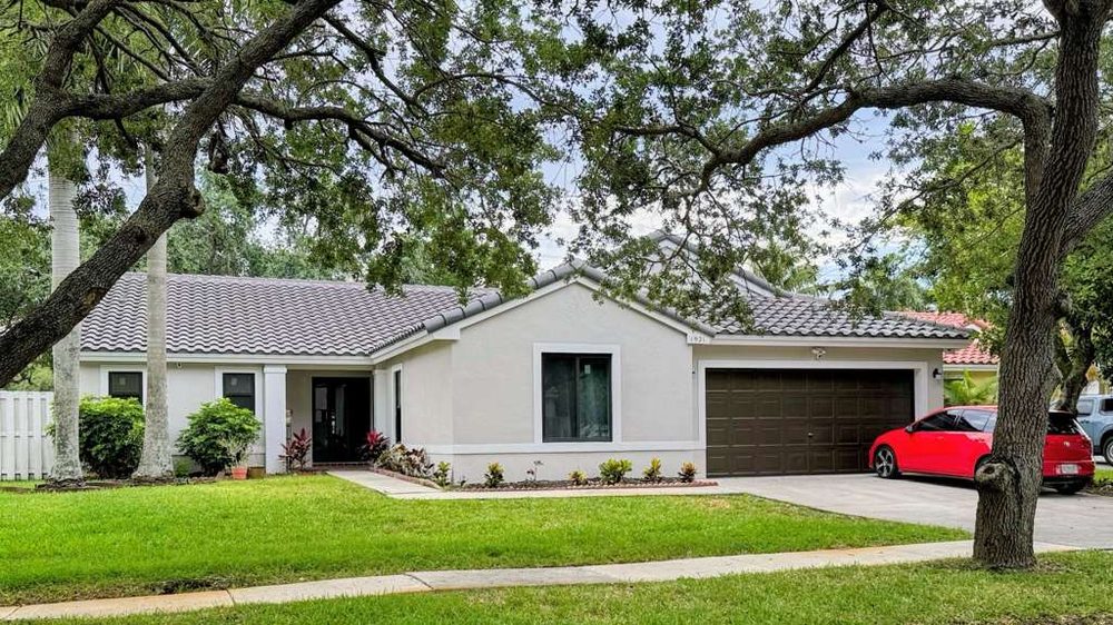 The same house after renovation: white stucco, a new grey tile roof, a dark bronze garage door, dark-framed windows and planted beds along the driveway.