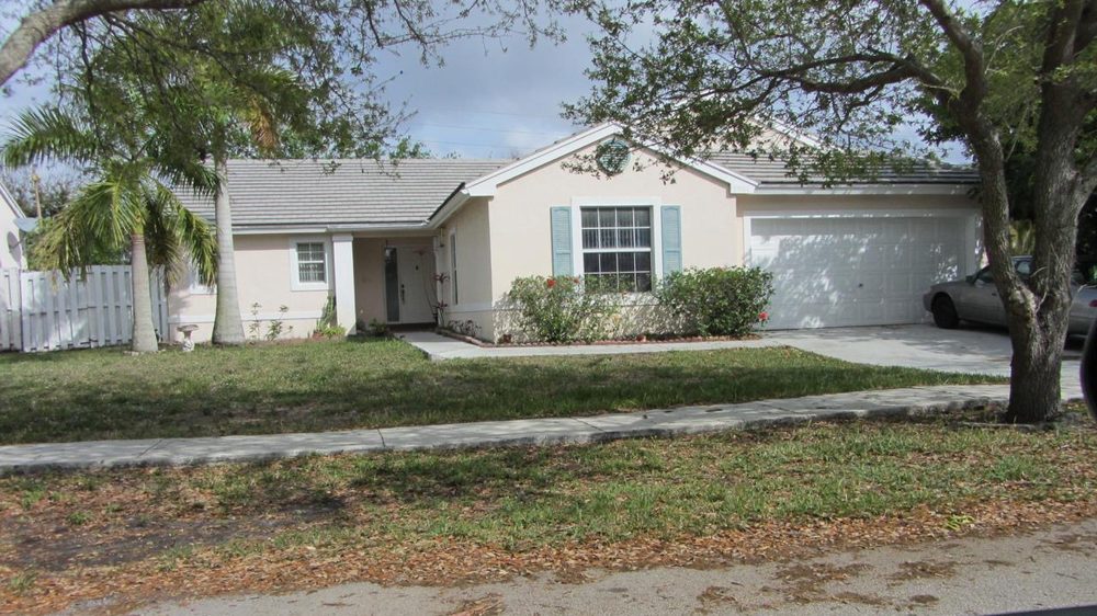 Single-storey South Florida house before renovation: beige stucco with teal shutters, an ageing tile roof, a plain white garage door and a patchy front lawn.